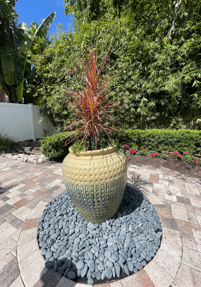 Residential landscaping installation - large light yellow and white round flower pot with tie sister plant sits on gray landscaping rock in the center of backyard pavers